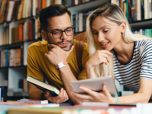 A young man and woman sit close together at a library table, contemplating a tablet the woman is holding up.