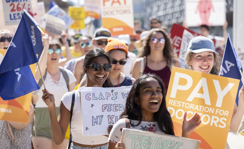 A group of smiling doctors march, hold handmade and BMA placards and wave flags in the sun outside at a junior doctor pay restoration march that took place on 15 June 2023